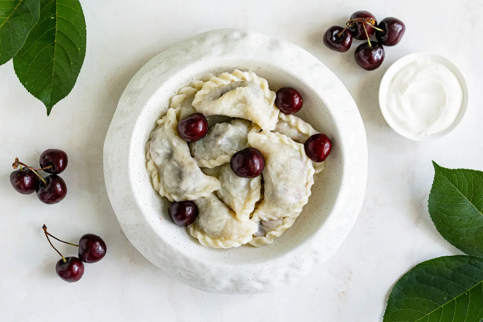 Varenyky in a stone bowl, surrounded by fresh cherries and cherry leaves